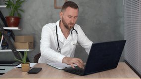 A young male doctor in a white medical coat sits in the office at the table and enters data from the table into his laptop.