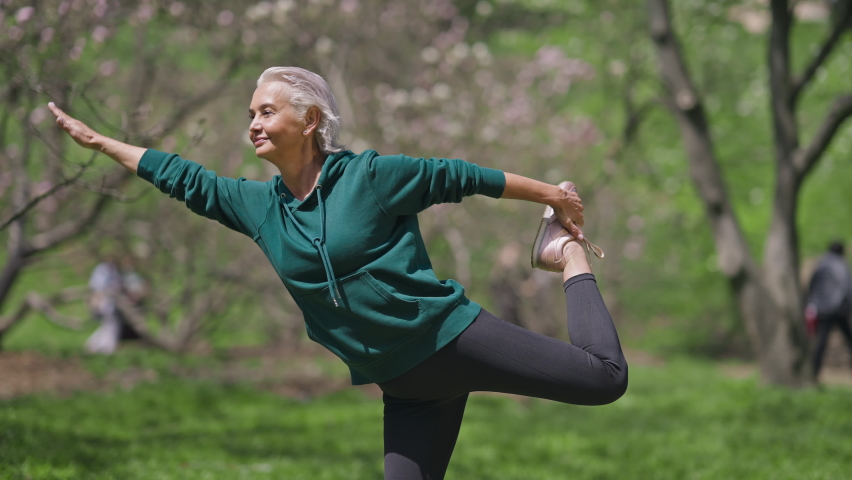 Happy senior sportswoman standing on one leg stretching muscles outdoors. Warmup exercise of positive confident fit Caucasian retiree in sunny spring summer park. Lifestyle and sport concept