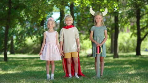Three children in costumes looking at camera and screaming with excitement in park. Boy wearing red cape and king crown and holding sword, girls in princess diadem and viking helmet. Kids role playing
