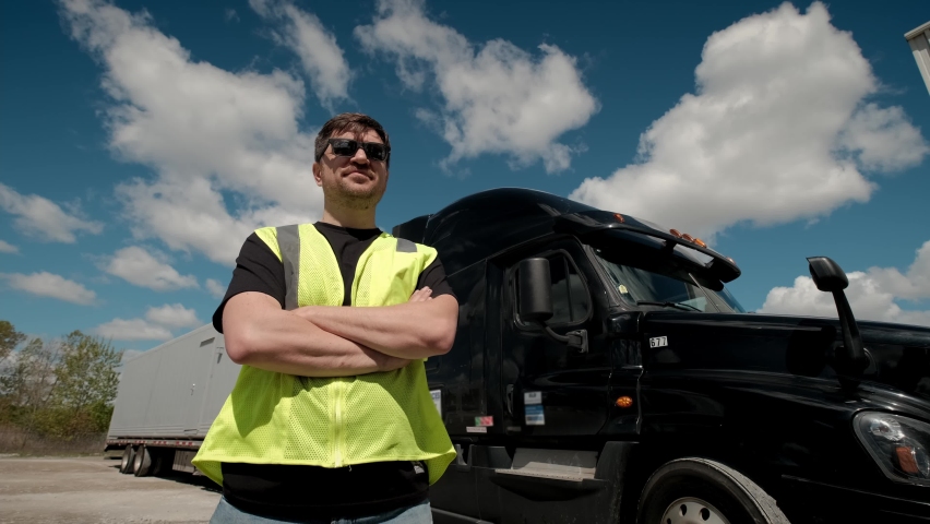 Professional Truck Driver in a yellow waistcoat approaches his truck and crosses his arms Behind Him Parked Long Haul Semi-Truck with Cargo Trailer Royalty-Free Stock Footage #1075822790