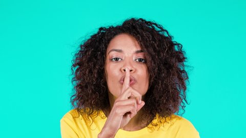 Smiling multi-ethnic woman holding finger on her lips over teal background. Gesture of shhh, secret, silence. Close up african lady..