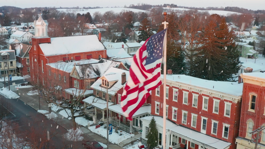 Small town America in winter. USA flag waves with snow flakes. Home, business, house apartment view. Aerial drone shot. Christmas holiday season.