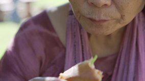 This close up video shows an old asian lady chewing and eating a Vietnamese sandwich outdoors.
