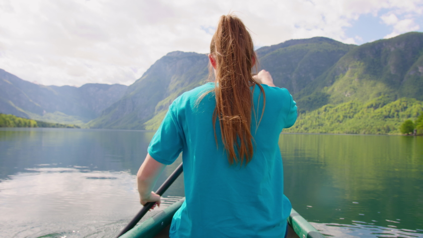 Handheld shot of a Caucasian young woman canoeing using a single-bladed paddle, traveling down a beautiful mountain lake, back view
