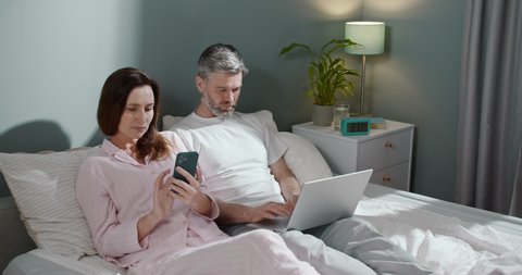 Beautiful happy caucasian adult couple relaxing on bed at home, man working on laptop computer while his cute wife reading something at the smartphone. People and gadgets concept