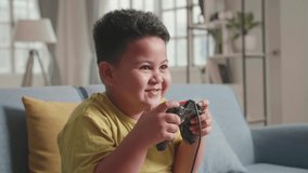 Young Asian Boy Using Joystick Playing Video Games In Living Room, Child Feeling Happy Using Relax Time Sitting On Sofa At Home
