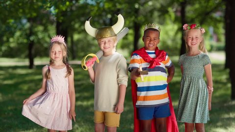 Group of diverse children in costumes looking at camera and screaming joyfully. Boys wearing red cape, king crown and viking helmet, girls in princess diadem and flower headband. Kids role playing