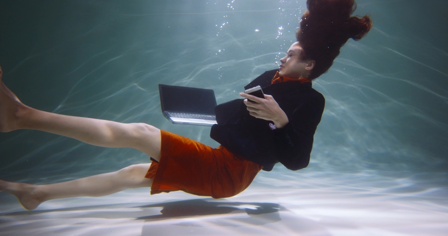 INFORMATION OVERLOAD. Cinematic side view, young business woman sinks under water with smart phone, laptop slow motion. | Shutterstock HD Video #1075837622