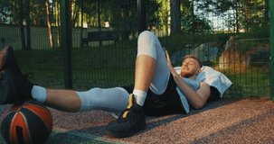 Basketball player sportive young guy lying with phone after workout outdoors on basketball court