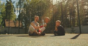 Male basketball player instructor training boy to spin a ball on finger on basketball court outdoors in sunlight