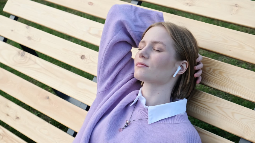 Relaxed young woman blonde in wireless headphones leans on brown wooden bench listening to music in park. Enjoying listening to lounge music live via smartphone app, calm without stress, eyes closed,