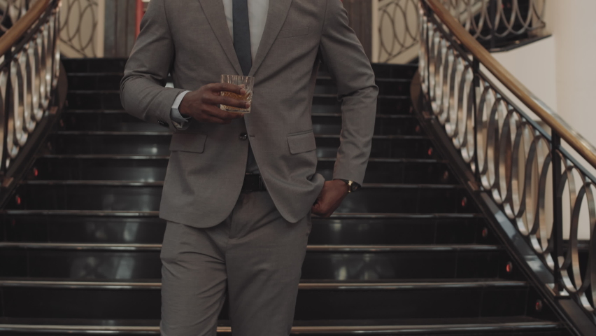 Tracking medium of African successful businessman holding whiskey glass, drinking, walking in premises with classy interior design