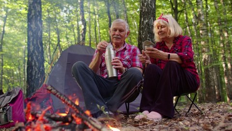 Senior elderly grandmother grandfather drinking tea over campfire in wood. Old man woman tourists having rest pouring hot drink from thermos at camping in forest. Active rest outdoors of mature family