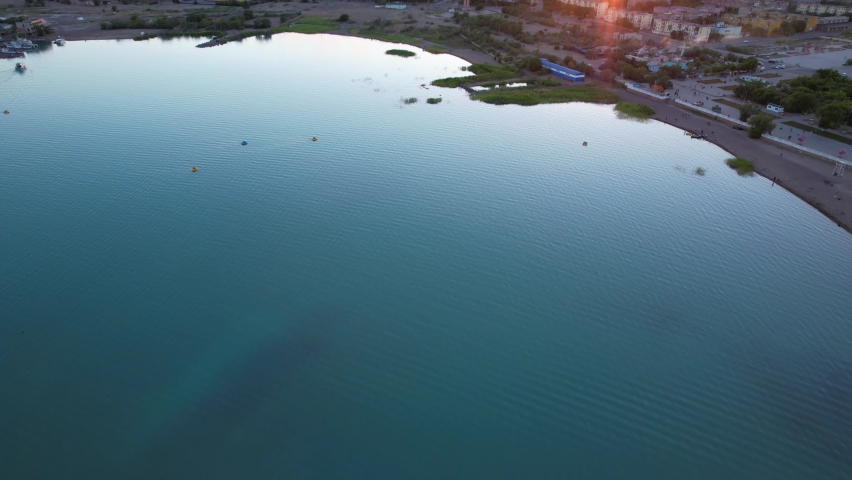 A lake, a factory and a city at a beautiful sunset. Catamarans and boats are floating on the lake, the sun is shining orange. A huge factory pollutes the air. Balkhash city beach. Kazakhstan