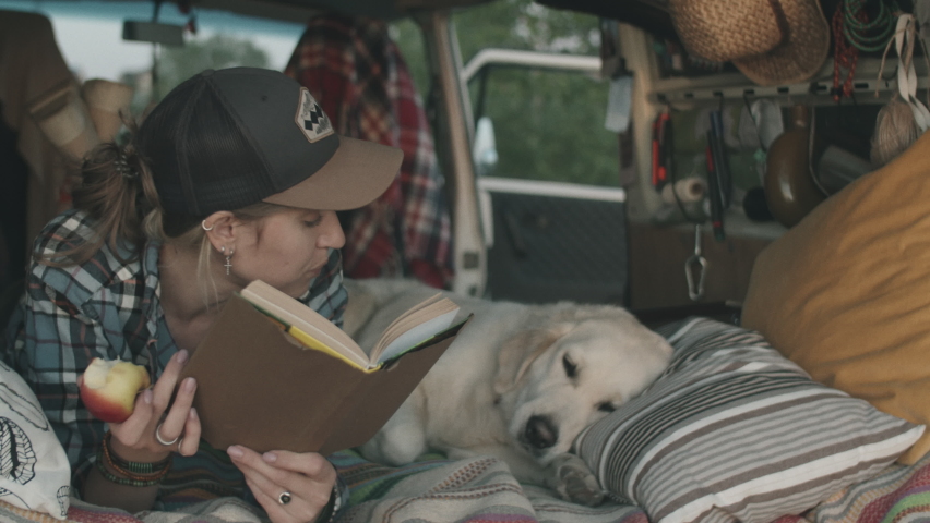 Young Caucasian woman lying in van, eating apple and reading book aloud to cute golden retriever resting near her
