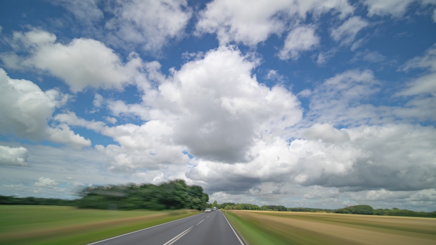Dashcam view. Driving on the narrow two-lane rural road with heavy oncoming traffic through the villages and farm fields. Thick white clouds above.