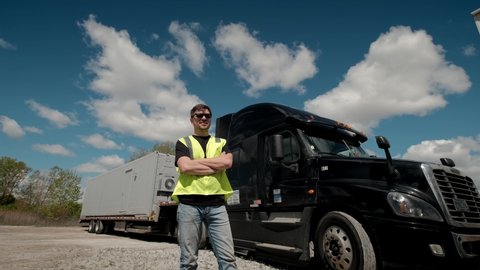 Professional Truck Driver in a yellow waistcoat approaches his truck and crosses his arms Behind Him Parked Long Haul Semi-Truck with Cargo Trailer