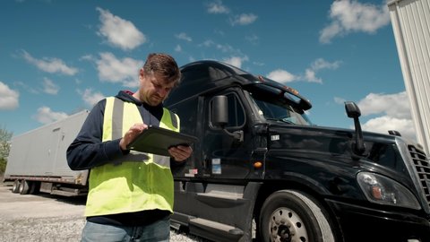 Truck driver standing by the truck in a yellow vest and using a tablet to fill a lookbook