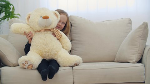 Small smiling girl sitting on the sofa with her toy teddy bear in 4k slowmotion video.