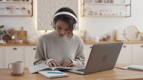 Young female student in wireless headphones passing exams during video call on modern laptop. Caucasian woman sitting at kitchen table with notes and books.