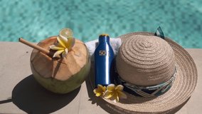 Video footage of green young coconut close up with bamboo straw, bottle of sunscreen, sun hat, tropical yellow flower frangipani, palm shade on the edge of swimming pool in Bali
