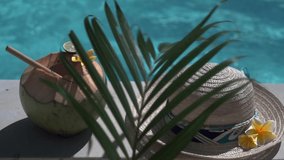 Video footage of green young coconut close up with bamboo straw, sun hat, tropical yellow flower frangipani, palm shade on the edge of swimming pool in Bali