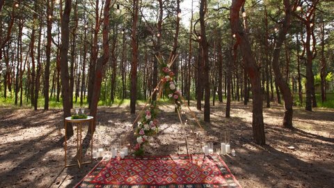 Beautiful bohemian tipi arch decoration on outdoor wedding ceremony venue in pine forest with cones. Chairs, floristic flower compositions of roses, carpet, string fairy lights. Summer rural wedding.