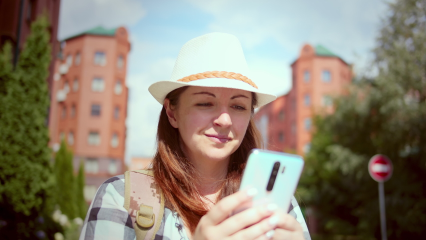 pretty woman in a hat chatting on the phone with friends against the background of red houses and smiling, woman in a good mood in summer, rack focus, cinematic shot