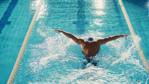 Successful Male Swimmer Racing, Swimming in Swimming Pool. Professional Athlete Determined to Win Championship using Butterfly Style. Colorful Cinematic Shot. Back View Aerial Tracking Slow Motion