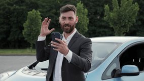 Positive businessman having video chat on smartphone near new electric car outside. Smiling professional call video online on cellphone business man using mobile phone outside Video conference.