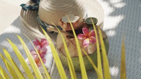 Video footage of green young coconut close up with bamboo straw, sun hat, tropical flowers frangipani, palm shade on white sun bed in Bali 