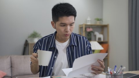 slow motion Japanese male returning to the working table is gulping tea in his hand and reading a paper mail bank statement in the living room at home.