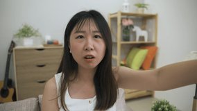 joyful asian young lady facing camera is touching her face and speaking with excited expression while enjoy distant communication with family at home.