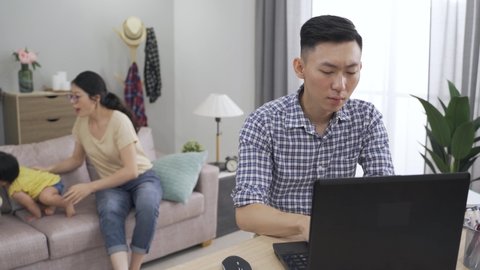 asian worker is mumbling with an annoyed look for his wife and toddler girl are making noise as he is typing a report on the laptop in the living room