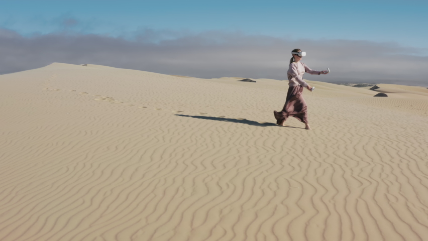 Girl uses virtual or augmented reality glasses in magic atmosphere of summer desert on sand dunes, female VR headset user on digital interactive art performance, entertainment of future. 4K aerial Royalty-Free Stock Footage #1075884266