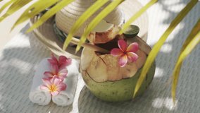 Video footage of green young coconut close up with bamboo straw, sun hat, tropical flowers frangipani, palm shade on white sun bed in Bali 