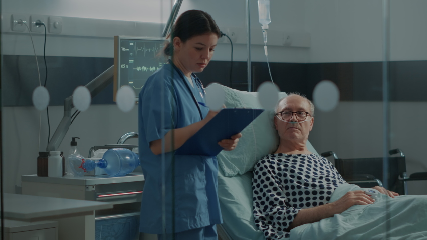 Caucasian nurse checking on elder patient in hospital ward at medical recovery unit. Old man with IV drip bag and nasal oxygen tube getting help to treat heavy health problems and illness Royalty-Free Stock Footage #1075885265