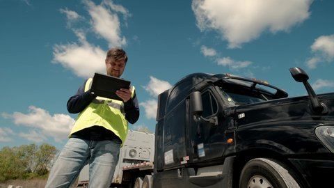Truck driver standing by the truck in a yellow vest and using a tablet to fill a lookbook. Wide shot