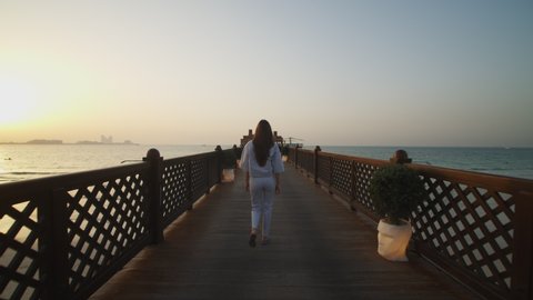 Woman walking on wooden pier and looking at Burj Al Arab hotel in Dubai. Back view of female tourist walking on wooden pier by sand beach in Dubai, UAE in 2021