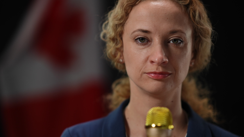 Close-up of confident journalist speaking in microphone at black background with Canadian flag. Portrait of serious elegant reporter talking telling news looking at camera