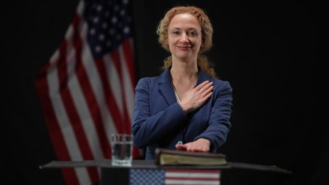 Confident elected politician woman holding hand on chest and on Bible looking at camera listening national anthem. Portrait of successful lady taking the oath at black background with American flag