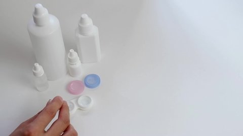 A female hand with tweezers places a contact lens in a plastic container on a white background with vials, bottles with eye drops. Daily lens care.