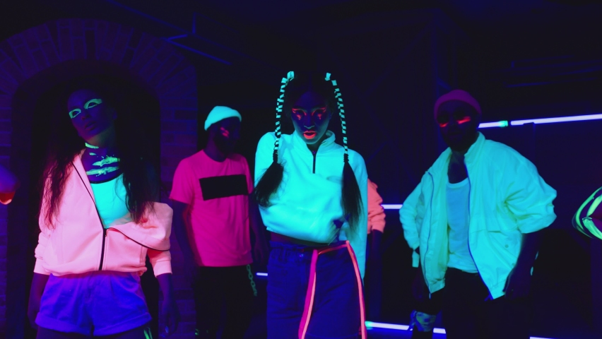 Group of female and male stylish dancers dancing inside dark place with neon lights .  Beautiful young people with fluorescent colorful make-up and clothes dancing in UV light. 4K video in slow motion