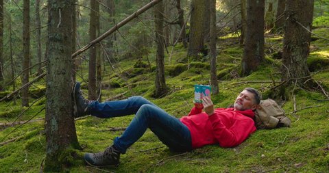 Outdoors travel concept. A bearded man is resting in a beautiful forest. He lies on the moss and reads a book. Slow motion. Ultra 4K
