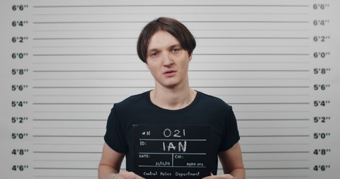 Profile mugshot of male person with dark hair turning to different sides and looking to camera. Criminal young man holding sign for photo while standing in front of police metric lineup wall