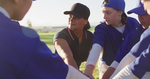 Diverse group of female baseball players and coach on pitch, in a huddle, stacking hands. female baseball team, sports training and game tactics.