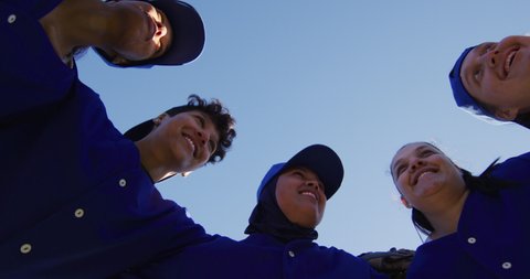Low angle view of diverse group of female baseball players in a huddle against blue sky. female baseball team, sports training and game tactics.