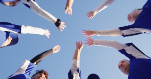 Low angle view of diverse group of female baseball players making hand stack against blue sky. female baseball team, sports training and game tactics.