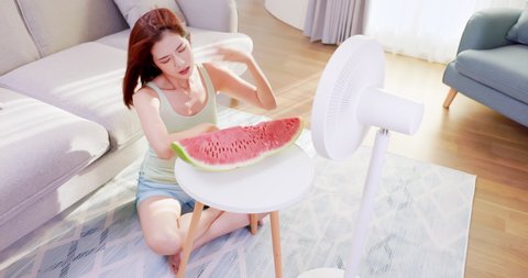 asian unhappy woman is sitting in the front of working fan with watermelon suffering from summer heat at home