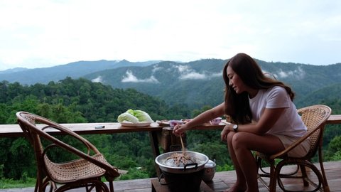 A woman cooking and eating Moo Kata, Thai barbecue grill pork together on balcony with beautiful nature view